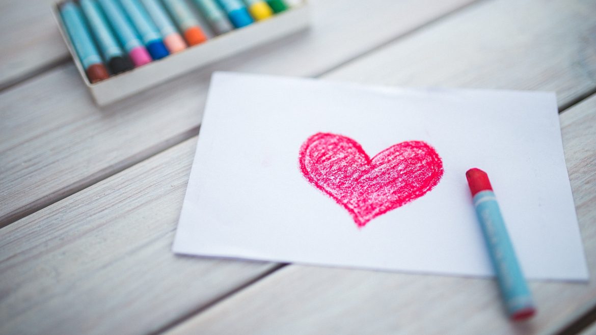 A piece of paper with a red heart drawn in pastel. Behind the paper there is a box of pastels waiting to be used.